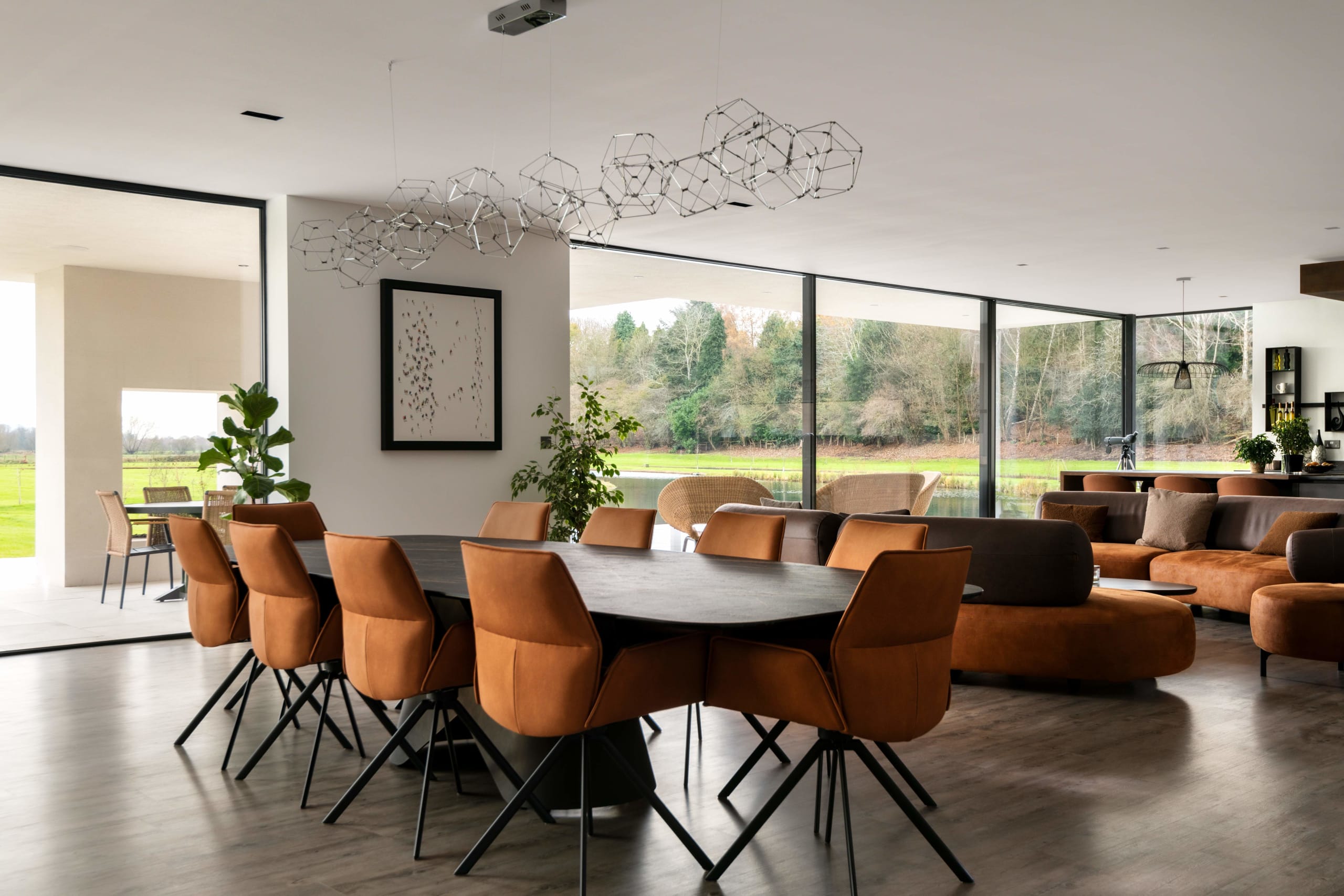 Modern open-plan living dining area with tan leather chairs, dark oval table, geometric chandelier and floor-to-ceiling windows